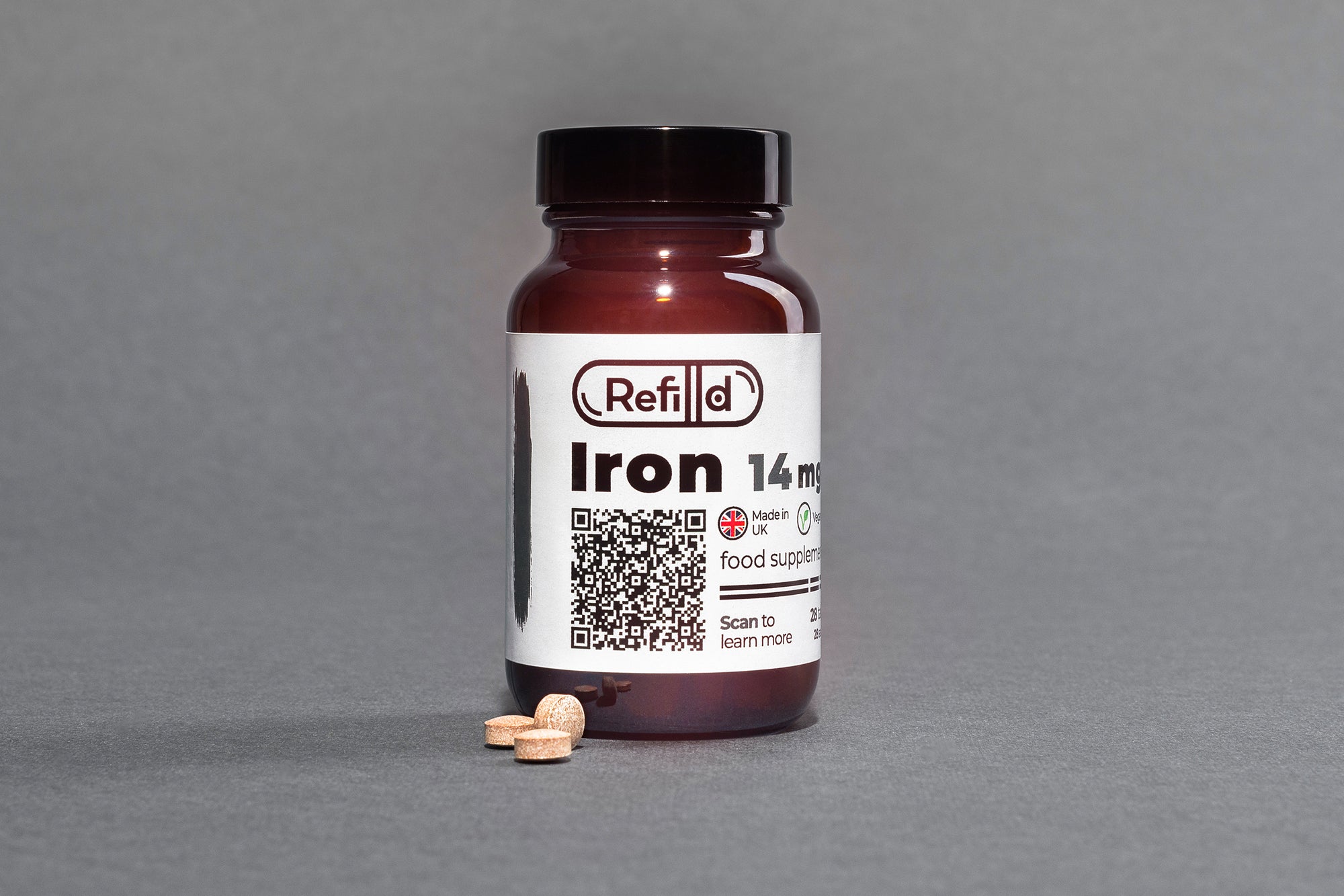 Refilld Iron 14 mg food supplement bottle with a few tablets in front on a gray background. Iron supplement, 14 mg, bottle, tablets.