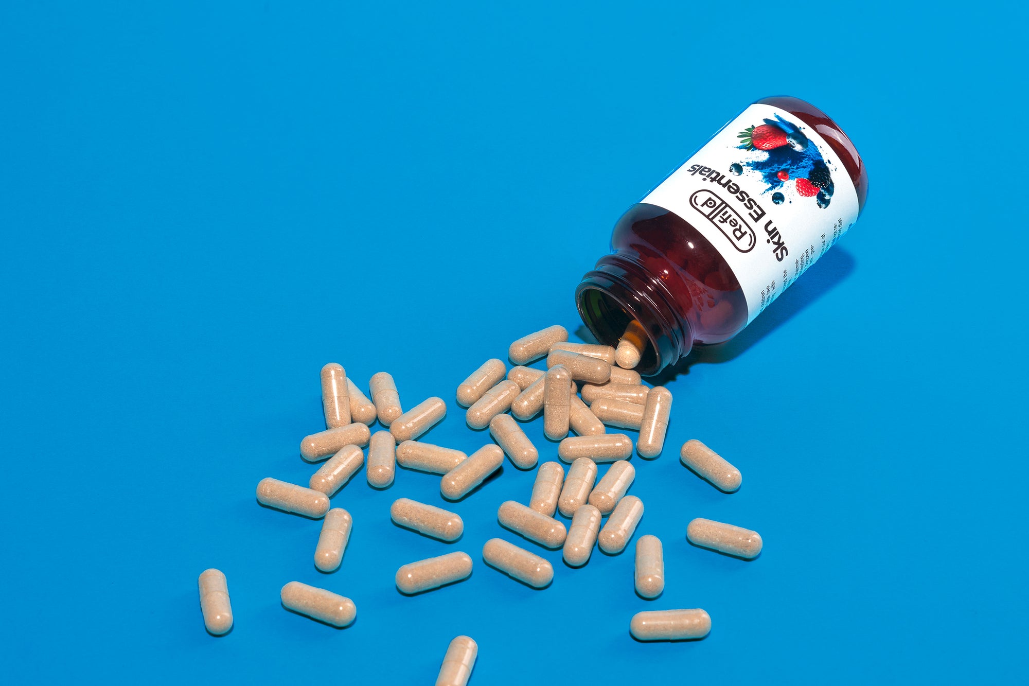 Spilled Refilld Skin Essentials bottle with beige capsules scattered on a blue background. Refilld Skin Essentials, food supplement, spilled capsules, skin health, blue background