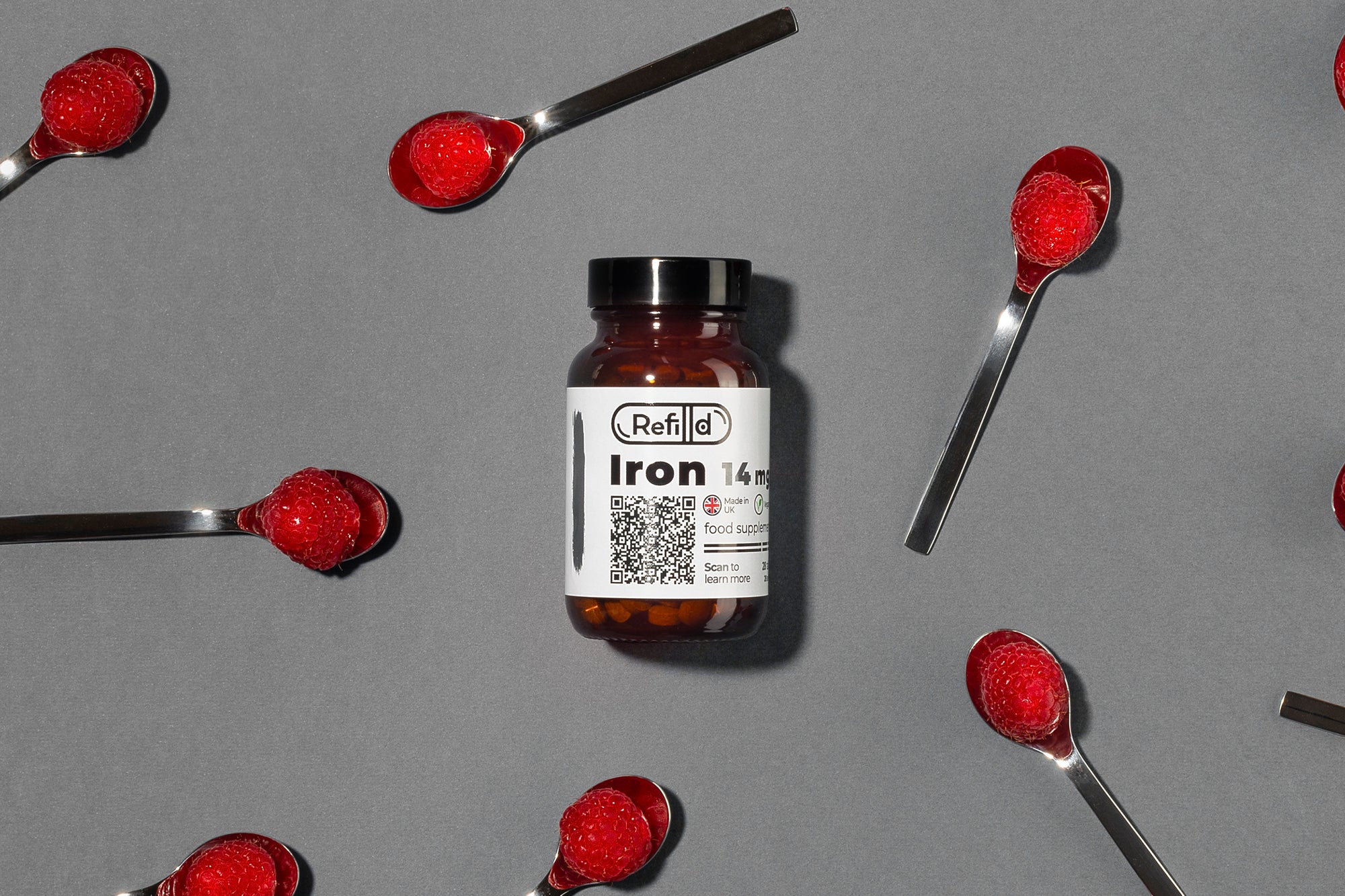 Refilld Iron 14 mg bottle with spoons holding raspberries on a grey background. Iron supplement, raspberries, iron-rich foods, gray background.