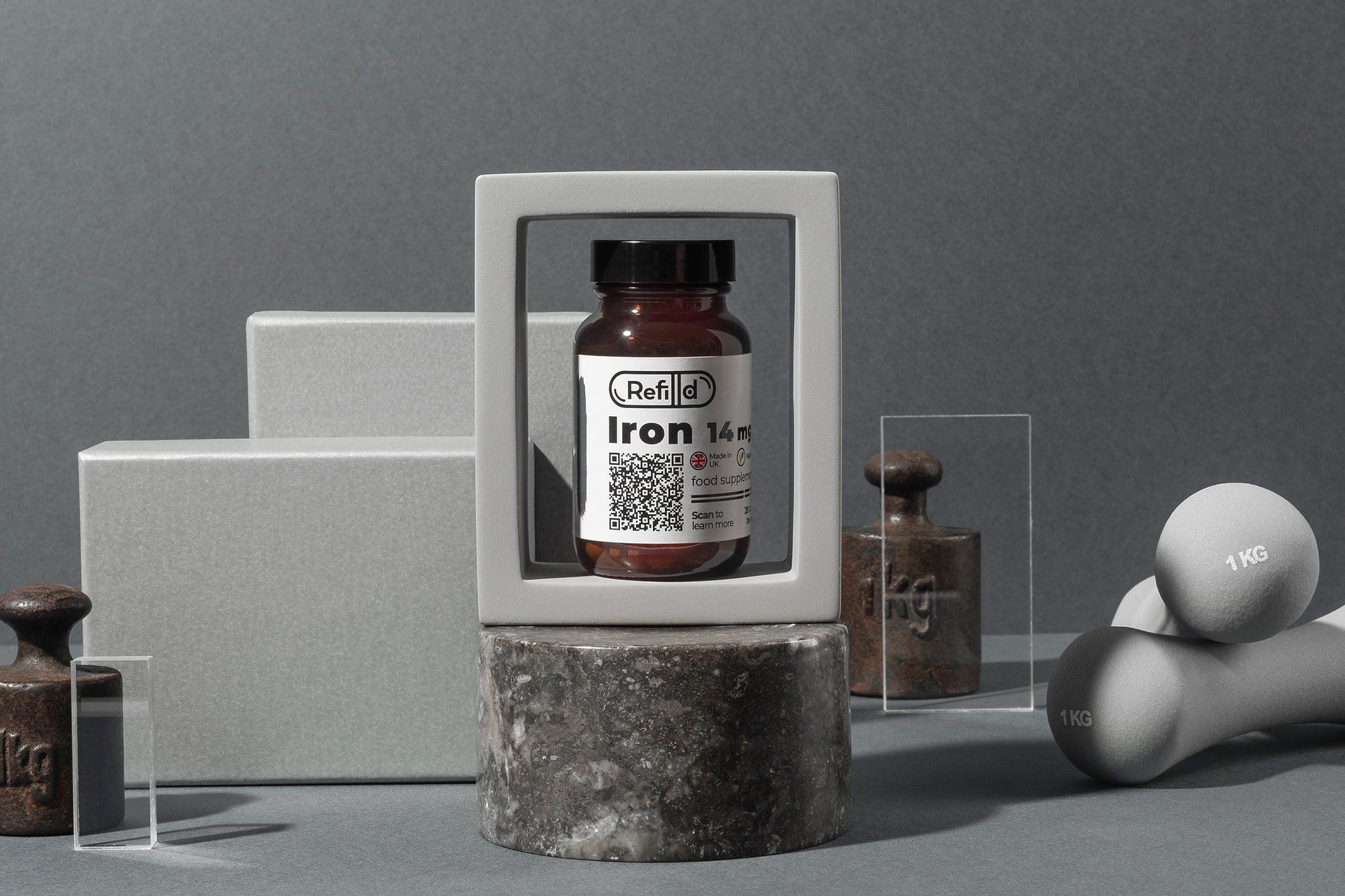 Refilld Iron 14 mg bottle with gym weights and dumbbells in a minimalist setup. Iron supplement, gym, fitness, minimalist setup.