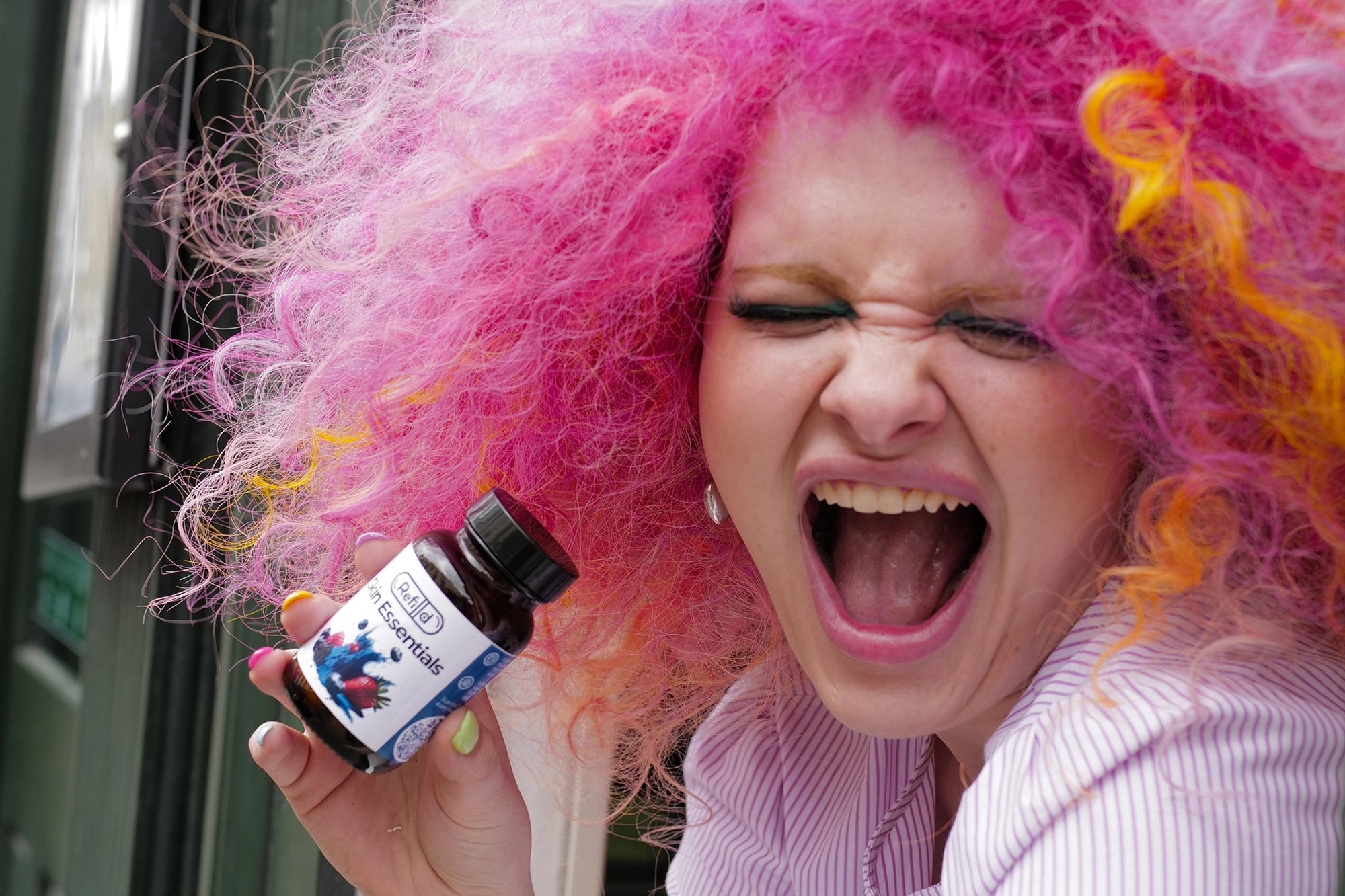 Rosie laughing and holding Refilld Skin Essentials bottle in hand, vibrant pink hair. Refilld Skin Essentials, skincare vitamins, fun brand character.