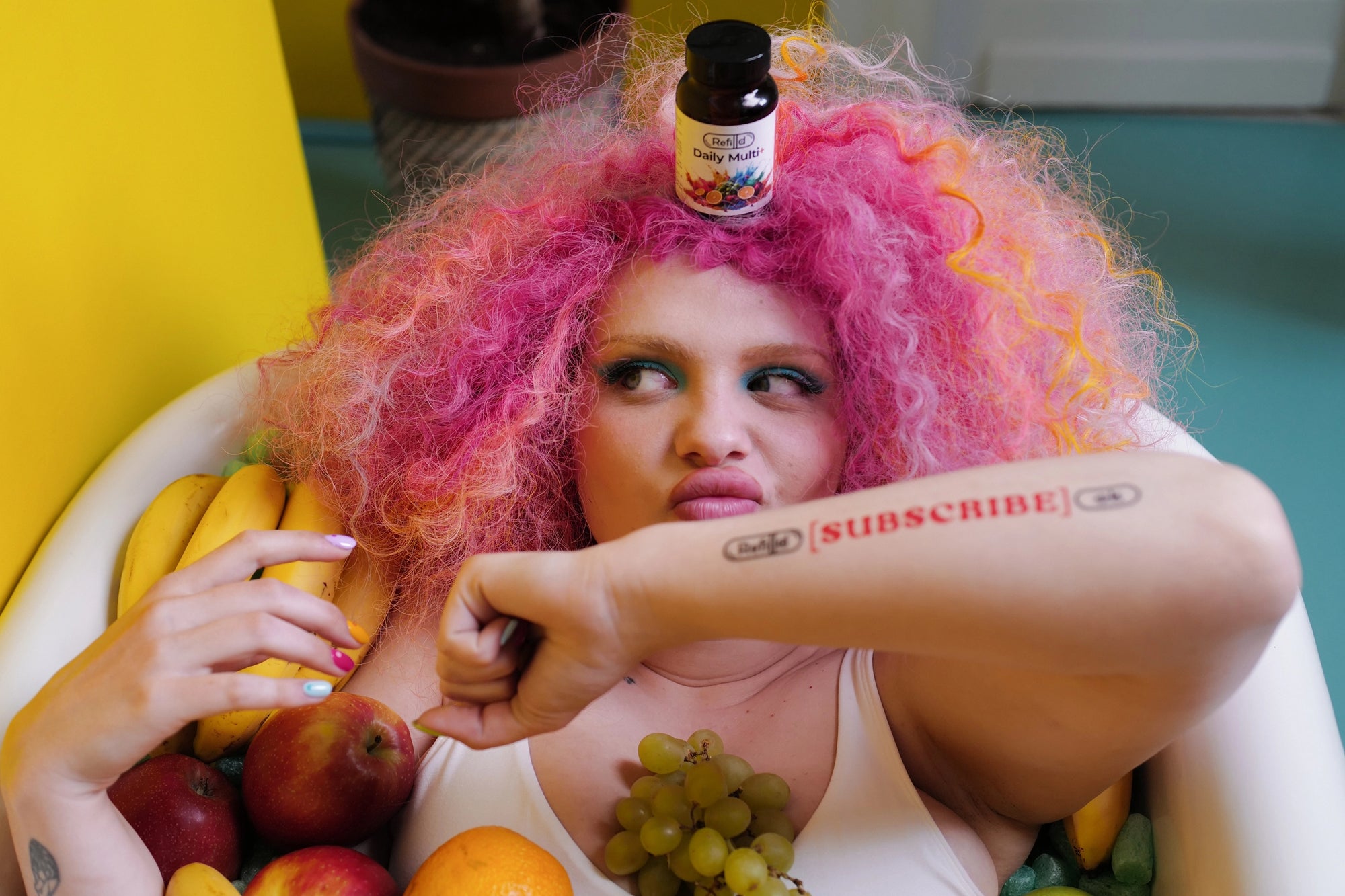 Rosie with Refilld Daily Multi+ bottle on her head, sitting in a tub filled with fruits. Subscribe to Refilld, vibrant pink hair, multivitamin supplement.