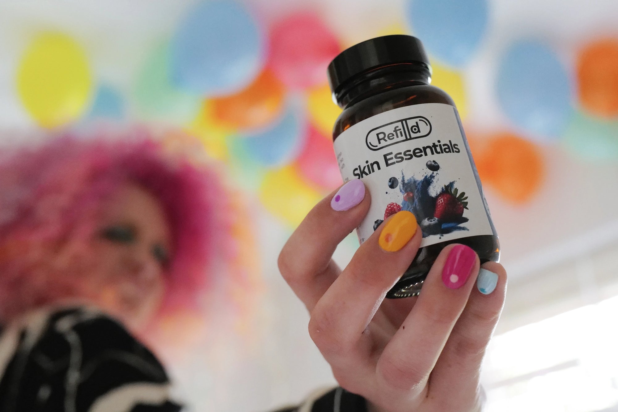 Rosie showing Refilld Skin Essentials bottle close to the camera, colourful nails, fun setting with balloons. Skincare vitamins, vibrant energy, Refilld marketing character.
